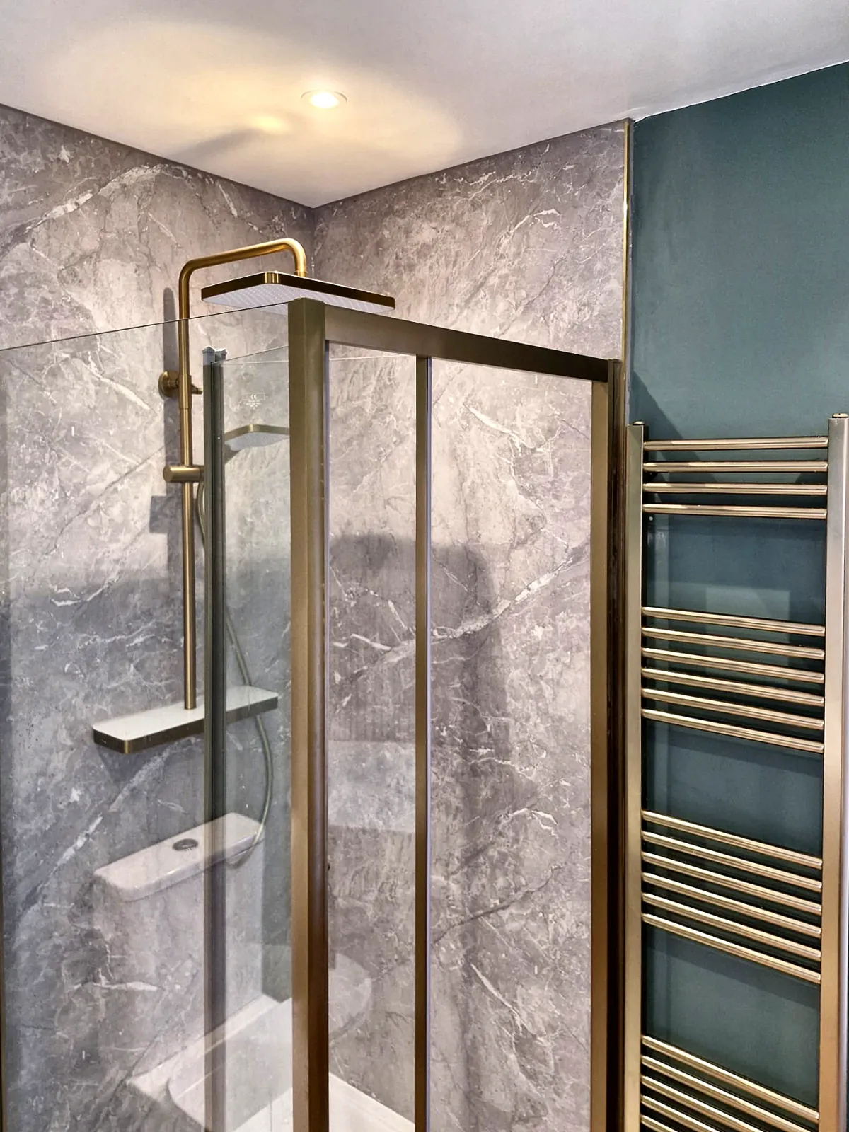 A shower enclosure with a brushed-brass frame and rain shower, grey marble-effect panels, and a brass heated towel rail beside it.