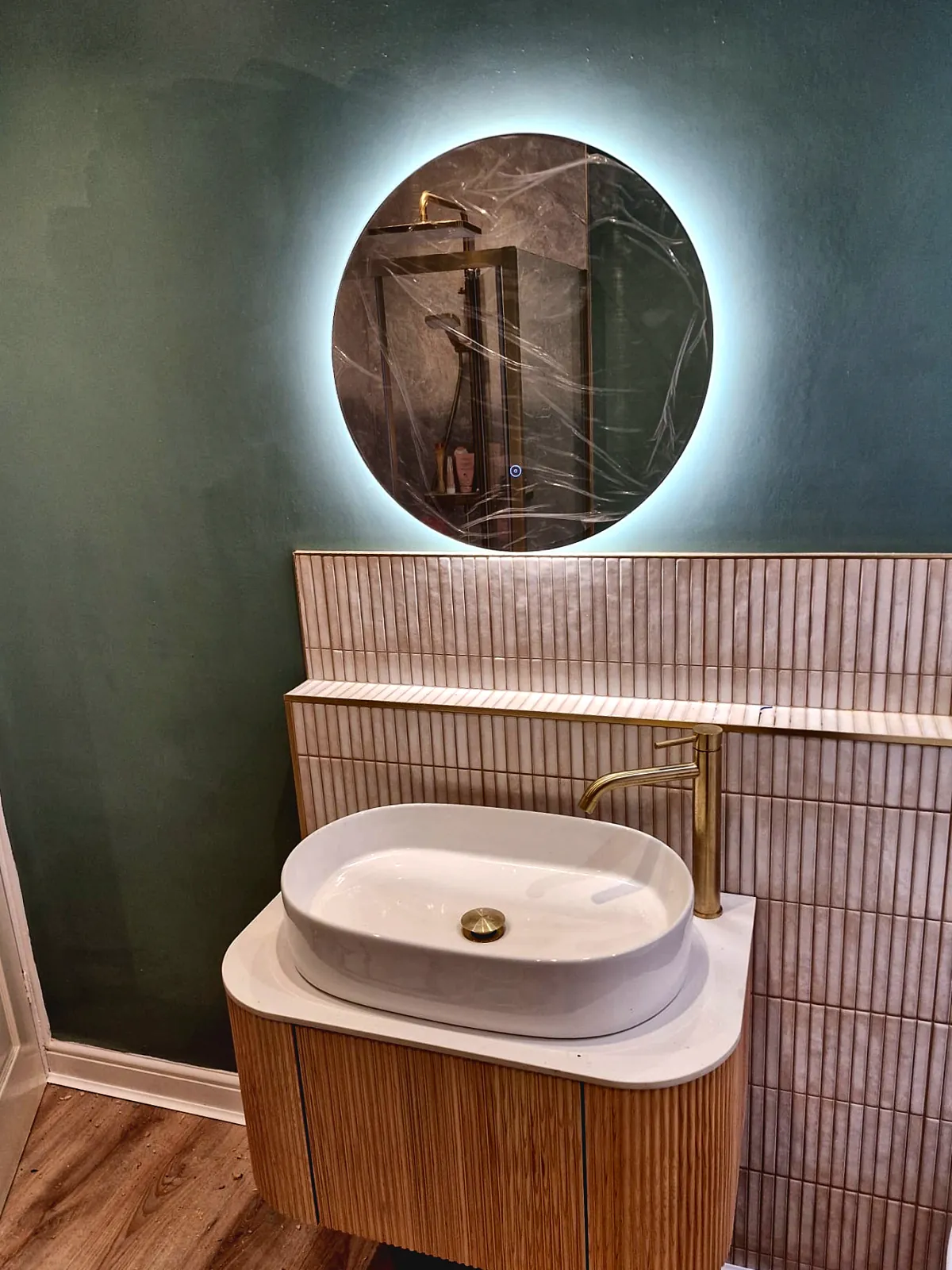 A wall-hung fluted wooden vanity with a white vessel basin and brushed-brass tap, under a round backlit mirror, against a green wall with fluted tiles.